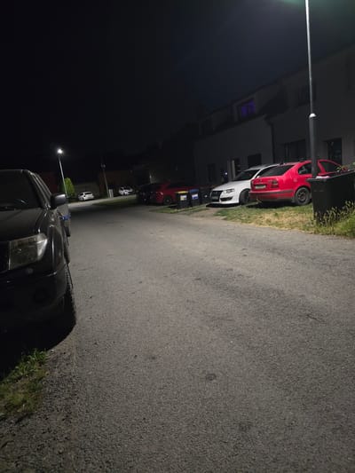 A nighttime street scene in Kostelec na Hané, Czechia, captures a parked black SUV in the foreground on the left, its headlights illuminated. Beyond the SUV, a paved road stretches into the background, lined with several parked cars. A white sedan is parked next to a red hatchback. Further down the road, two white compact cars are visible. A streetlamp casts a bright light over the scene. Buildings with illuminated windows are visible in the background. The environment appears to be a residential area.