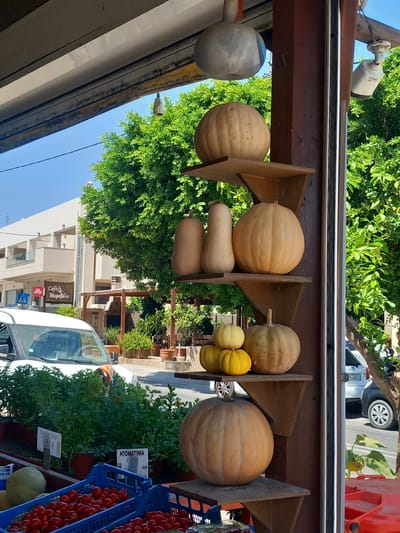 The image shows a market stall display of pumpkins and other produce in Platani, Greece. A large, light-colored pumpkin is prominently featured on a wooden shelf, with several smaller pumpkins and gourds stacked below it. To the left, a white van is parked, and in the background, a building with balconies and trees can be seen. The scene appears to be outdoors, with sunlight illuminating the display. The overall impression is one of a local market in a Mediterranean setting.