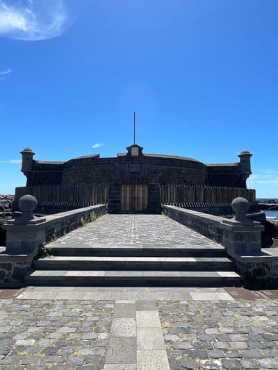 The image shows the Castillo de San Juan Bautista, also known as the Black Castle, in Santa Cruz de Tenerife, Spain. It is oriented vertically with the castle in the center. The fort is constructed of dark stone and has a simple, fortress-like design with a central body and two cylindrical turrets with domes on either side. In front of the castle, there is a stone pathway with steps that lead to the entrance. The path is made of cobblestones and stone slabs. The sky is clear and blue, with a few wispy clouds visible. The ocean is visible in the top right corner of the image. The structure appears to be well-maintained, and no people are visible in the scene.