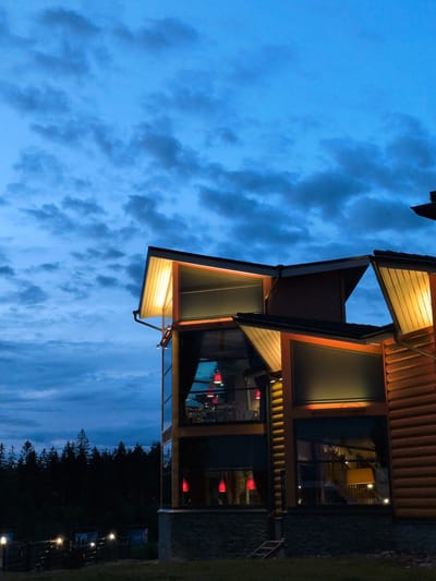 This is a photograph of a modern wooden building with large windows, taken at dusk. The sky is a deep blue with scattered clouds. The building's exterior is lit by warm, yellow interior lights and orange exterior accent lighting. Red pendant lights are visible hanging inside through the windows. To the left of the building, a dark forest of evergreen trees is visible against the sky. In the foreground, there is a grassy area and some fencing with several illuminated lamps. The image is not specifically located in Heinola, Finland.