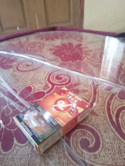 A pack of "Apel 76" cigarettes, a Indonesian brand, lies on a red patterned tablecloth. The pack is partially covered by a clear plastic sheet. The cigarettes are filter-tipped and the pack features warnings about the dangers of smoking. The background shows a wooden surface, possibly a table or wall. The scene is likely indoors, given the tablecloth and the general ambiance. The image does not provide any specific information to determine its exact location within Sukamanah, Indonesia.