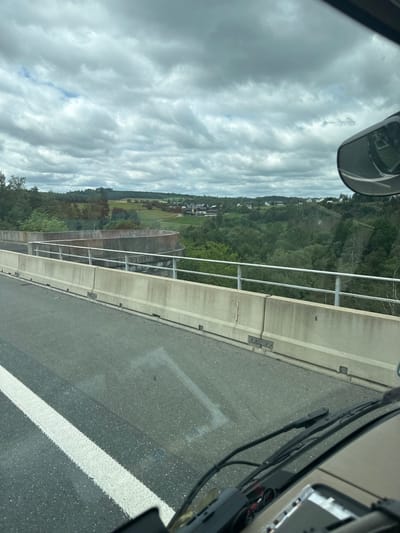 The image is taken from the perspective of a vehicle driving on a highway overpass in Köditz, Germany. The highway has a grey asphalt surface with a white lane marking on the left side. The overpass is bordered by concrete barriers and a metal railing. Beyond the overpass, a lush green valley with scattered houses and buildings is visible. The sky is overcast with dramatic grey clouds. The overall scene depicts a rural landscape in Germany seen from a moving vehicle.