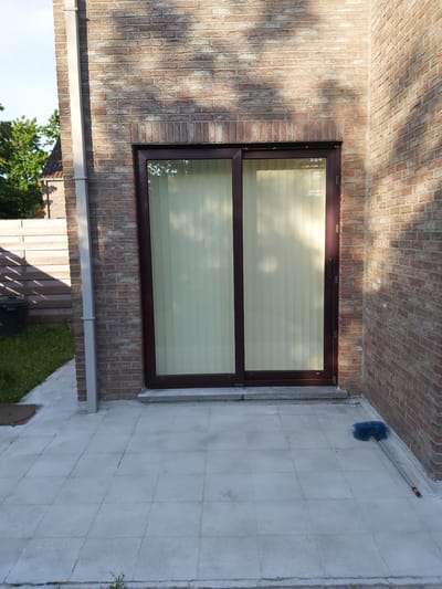 This is a ground-level exterior, eye-level, medium shot of a sliding glass door on the side of a brick building in Dendermonde, Belgium. The door has a dark brown frame and vertical blinds behind the glass. The brickwork is reddish-brown with some lighter-colored mortar. To the left of the door is a downspout, and to the right is the corner of the building. The ground in front of the door is paved with light gray square tiles. A blue-headed broom with a long handle lies on the tiled patio to the right of the door. In the background to the left, there is a wooden fence and some greenery. The sky is visible at the top left.