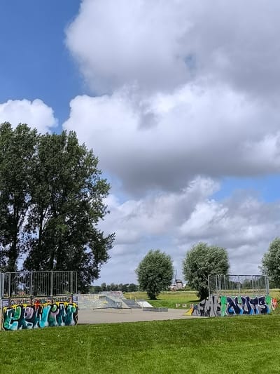 The image displays an outdoor skate park situated within a broad, grassy expanse under a dynamic sky. The scene is illuminated by bright daylight, with patches of clear blue sky interspersed among large, white and grey cumulus clouds, indicating partially cloudy weather.
The skate park features various concrete ramps and rails. In the foreground, on both the left and right, are concrete structures serving as barriers or ledges, extensively covered in graffiti. On the left structure, prominent green, blue, and yellow letters spell "WOKE," with smaller, less distinct text above it that appears to include "RISE" and "DAK." On the right structure, large blue and black letters clearly read "NUTS," accompanied by an adjacent, illegible black and white graffiti design. Both structures are topped with metal railings.
No individuals are visible in the skate park, but a red bicycle is leaning against the right graffiti wall, suggesting recent presence or anticipated use. The immediate foreground consists of vibrant green grass, which also surrounds the entire skate park area. Tall, leafy trees frame the left side of the image, and several other round-canopied trees are dotted across the middle ground. In the distance, beyond the trees and fields, a classic Dutch windmill with a white body and dark cap is discernible, establishing a strong location cue for the Netherlands, specifically Oudorp. The overall impression is one of a spacious, semi-rural recreational area.