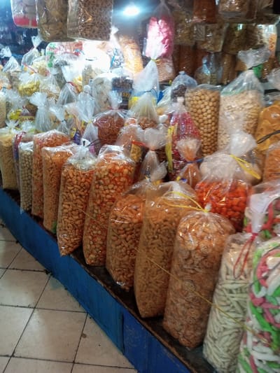 This indoor market stall or shop, located in Lê Thanh Nghị Ward, Vietnam, displays a dense arrangement of packaged snack foods. Numerous clear plastic bags, varying in size, contain diverse snack types including puffed items (light brown, white, orange/red), nuts, ring-shaped snacks, white stick-like crackers, and multi-colored intertwined items (pink, green, white). Each bag is sealed with a colored string or rubber band (red, yellow, blue, white). Many bags are stacked on a blue-painted wooden or metal display counter. The floor is composed of light-colored square tiles. Artificial overhead lighting illuminates the merchandise.