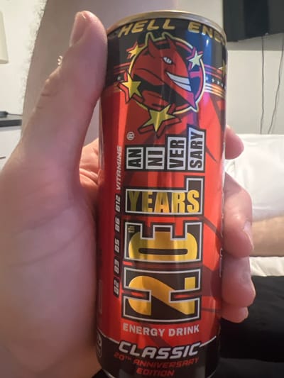 A human hand is holding a can of "HELL ENERGY DRINK CLASSIC 20TH ANNIVERSARY EDITION" with a red devil logo on a black background. The can is primarily red with black and gold accents. The words "ANNIVERSARY" and "YEARS" are prominently displayed. Text on the can also includes "ENERGY DRINK", the word "VITAMINS" and the letters "B2, B3, B5, B6, B12". The background includes a white sheet and dark clothing. There is no indication of the image's location within the city of Sheffield, United Kingdom, or any interaction taking place.