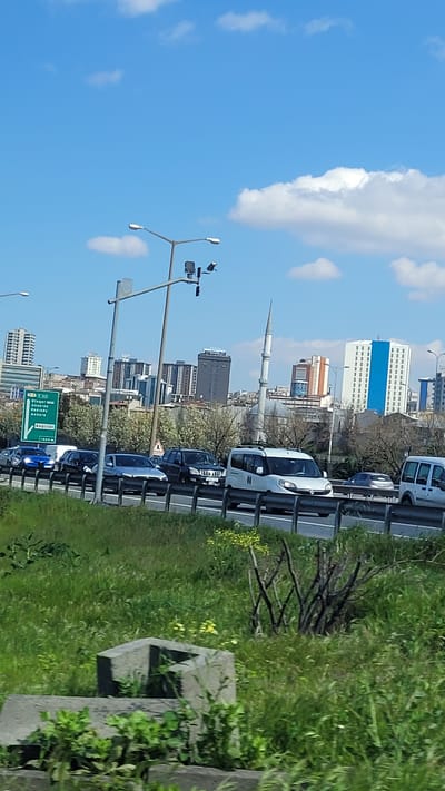 The image captures a view in Bağcılar, Turkey, likely taken from a moving vehicle. It shows an urban environment with a highway running alongside grassy terrain on the right side. Several vehicles are present on the highway, including cars and vans. In the background, multiple buildings are visible, including a mosque with a prominent minaret. Street lamps and road signs are positioned along the highway. The sky is blue with scattered clouds.