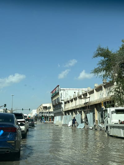 The image depicts a flooded street in Sharjah, United Arab Emirates. Cars are partially submerged in floodwater, indicating significant rainfall or water overflow. One individual is wading through the water near a building under construction, which has corrugated metal barriers. Traffic lights and street lamps are visible, along with commercial buildings and signage, some of which are for "A. ALICO." The sky is partially obscured by a tree in the upper left corner and features a combination of clear blue and scattered clouds.