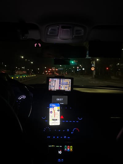 A car interior at night shows the dashboard illuminated with blue lights. A phone displays a map, indicating 6.2 km to the destination with an estimated arrival time of 06:53. Another GPS device shows a map as well, reading 06:37. Outside, traffic lights are visible, with red and green lights in the distance. Streetlights line the road. The car's interior light fixture is visible in the upper left corner.