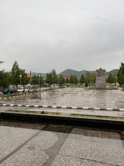 The image captures a cloudy day in Voiniagovo, Bulgaria, featuring a stone-paved public space. A monument of a man stands at the center of the square, framed by trees and mountains in the background. Street lamps line the walkway, and parked vehicles are visible near the trees. The foreground shows a concrete structure filled with water reflecting the surrounding environment.