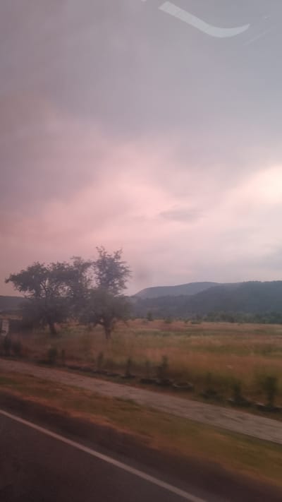 The image depicts a scenic landscape with a road running alongside a field and a mountainous backdrop. The sky is filled with soft, diffused light, hinting at either dawn or dusk, with subtle pink and purple hues blending into a pale grey. This atmospheric lighting creates a serene and somewhat melancholic mood.

In the foreground, a section of what appears to be a window or a reflective surface is visible on the left, with a curved white line suggesting a car's interior or framing. To the right of this, the sky dominates, with some cloud formations adding texture.

The middle ground features a field, predominantly in shades of brown and green, suggesting dry grass or harvested crops, with scattered trees. One prominent tree with dense foliage stands out. Beyond the field, a range of mountains stretches across the horizon, their slopes covered in dark green vegetation, indicating a forested area.

The right side of the image is occupied by a road, dark grey in color, with a white lane marking running along its edge. The road appears to be wet, possibly from recent rain, reflecting the muted light. The overall scene suggests a rural setting in Dubrynychi, Ukraine, during a tranquil period of the day, possibly after a rain shower, given the wet road. There is no visible text in the image.