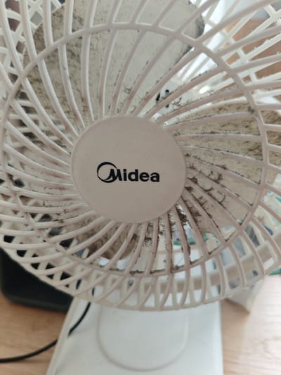 A close-up, slightly angled shot shows a dirty white Midea brand fan. The fan's grill, made of white plastic bars, is heavily covered in dust and debris, creating a gray and brown appearance. The Midea logo, a black stylized "M" within a circle, is visible on the center of the fan's protective grill. The fan is positioned on a white base, and a black power cord extends out of frame. The background suggests an indoor setting with a wooden surface visible. The image is taken from a perspective that emphasizes the fan's front. There are no people visible in the image. This image is located in Berdsk municipality, Russia.