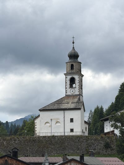 The image shows a white church with a prominent bell tower, located in the town of Sauris / Zahre, Italy. The church has a gray tiled roof and a tall, dark steeple topped with a cross. A clock face is visible on the bell tower. The church is situated on a raised stone wall, with a wrought-iron fence running along the top. Behind the church, there are lush green trees and mountains in the distance, partially obscured by a cloudy, overcast sky. In the foreground, wooden buildings with dark roofs are visible. The overall scene depicts a rural, mountainous setting under a moody sky.