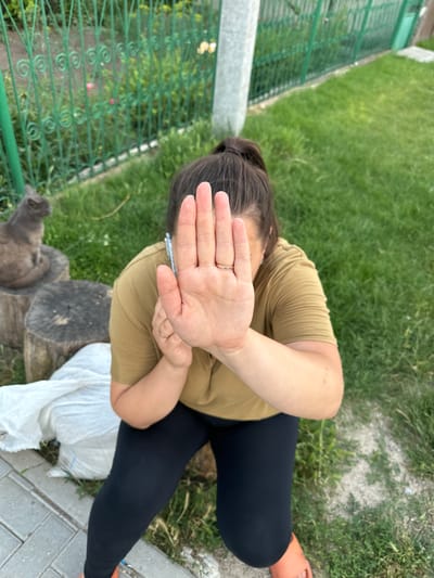 In this image, a person is sitting on the ground outdoors, in what appears to be a backyard or garden area. The person is wearing a tan t-shirt and black leggings, with orange footwear visible on their right foot. They are shielding their face with their right hand, suggesting an attempt to avoid being photographed. A ring is visible on the person's left ring finger.
The setting is grassy with some patches of dirt and paving stones. A green metal fence runs along the left side of the frame, with some plants visible beyond it. Two wooden stumps are situated in front of the person. A greyish-brown animal, possibly a cat or small dog, is partially visible in the lower left corner of the image, looking towards the person. A white plastic bag lies on the ground near the person's legs.
The overall impression is of a casual, perhaps spontaneous moment outdoors, with the subject attempting to obscure their identity. The lighting suggests it is daytime, and the lush green grass implies favorable weather conditions. The presence of the fence and garden elements point towards a residential setting in Pîrlița, Moldova. No text is visible in the image.
