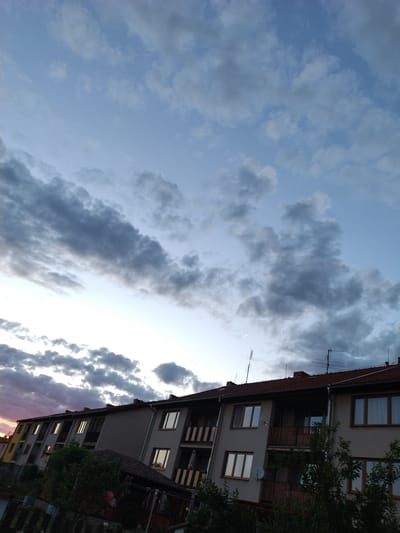 The image shows a view of apartment buildings against a dusky sky with clouds. The buildings are multi-story with balconies and windows, and some have satellite dishes and antennas on their roofs. Trees and a fence are visible in the foreground. The sky is a mix of blue and gray clouds, with hints of pink on the left side, suggesting sunset or sunrise. The perspective is slightly tilted, looking up towards the sky. The scene appears to be in a residential area.