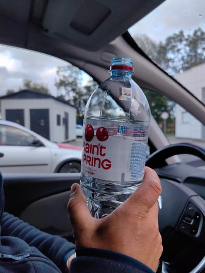 A first-person perspective shot shows a person's hand holding a bottle of Saint Spring bottled water inside a car. The bottle is partially filled and features a label with red text and two cherry graphics. The background, visible through the car windows, shows a cloudy sky and some trees, with a white building that appears to be a garage or small structure in the distance. The steering wheel of the car is also visible on the right side of the frame. The scene appears to be located in the vicinity of Фаніпальскі сельскі Савет, Belarus, given the Cyrillic text on the bottle's label, which includes production and contact information for a Belarusian entity.