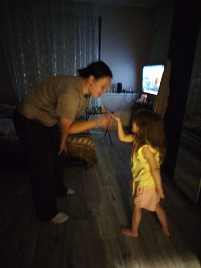 In a dimly lit room, an adult and a young child are engaged in an activity. The adult, with dark hair pulled back, is wearing a taupe t-shirt and dark pants. They are bent over, extending their right arm towards the child, with their hand open. The child, with long brown hair, is wearing a yellow sleeveless top and a light pink skirt. They are standing facing the adult, with their left arm raised and hand open, mirroring the adult's gesture. The room has a wooden floor. In the background, a television screen is illuminated, and there is a curtain on the left side of the frame. A rack with what appears to be clothing is visible on the right. The scene appears to be indoors, possibly a living space. There is no information within the image to suggest a specific location in the city of Krasny Gulyai, Russia.