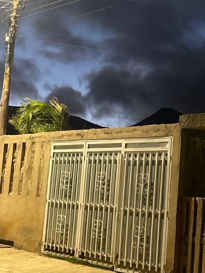 An outdoor, medium shot captures a foreboding sky over a low-lying structure in San Antonio, Venezuela. The sky is a dark, bruised blue with ominous, swirling gray clouds, suggesting an approaching storm. The top of a dark, conical mountain is visible against the sky. In the foreground, a roughly textured, tan-colored wall runs horizontally across the frame. Palm fronds, tinged green and yellow, are visible above the wall on the left, appearing windswept.

Set into the wall is a white, double-door structure with vertical bars. These bars are painted white and feature decorative, heart-shaped metalwork within them. Behind the bars, a dark, possibly reflective surface is visible, hinting at windows or glass doors. A weathered, wooden utility pole stands to the left of the wall, adorned with various electrical boxes and wires that crisscross the frame against the turbulent sky. To the right of the double doors, a section of weathered wooden fence is visible. The ground in front of the wall appears to be a paved surface, illuminated by artificial light, with a narrow strip of green grass at the base of the barred doors. The overall scene conveys a sense of unease and impending weather.