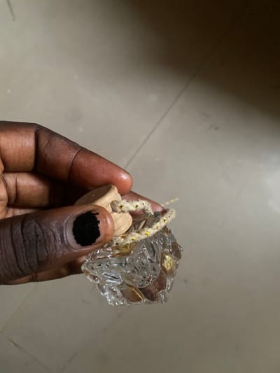 A close-up shot shows a person's hand holding a small, cube-shaped object. The object appears to be made of clear, faceted material, possibly glass or crystal, with small, metallic squares, some gold and some possibly brass or copper, embedded within it. A rough, natural fiber rope, with subtle gold threads woven through it, is attached to the object and also held by the person's fingers. The rope is tied to a small, light-colored wooden piece that is partially visible.

The hand holding the object is dark-skinned, with prominent fingerprints visible on the index finger and thumb. One fingernail, likely the index finger's, is painted black in a rough, abstract pattern. The background is a blurry, neutral-toned surface that suggests a floor, with subtle variations in light and shadow. A faint line runs diagonally across the background, which might indicate a seam or crack in the floor.

The lighting is somewhat dim, casting shadows and highlighting the reflective surfaces of the object. The overall impression is one of a personal, close-up examination of a decorative or possibly functional item. No specific time of day or weather conditions can be inferred. The setting appears to be indoors, given the floor surface and lighting, but no other environmental cues are present. The emotion conveyed is neutral, focused on the tactile interaction with the object. There is no visible text in the image. The context of Yola, Nigeria, does not provide specific visual cues within the image itself, but serves as a geographic location for the scene.