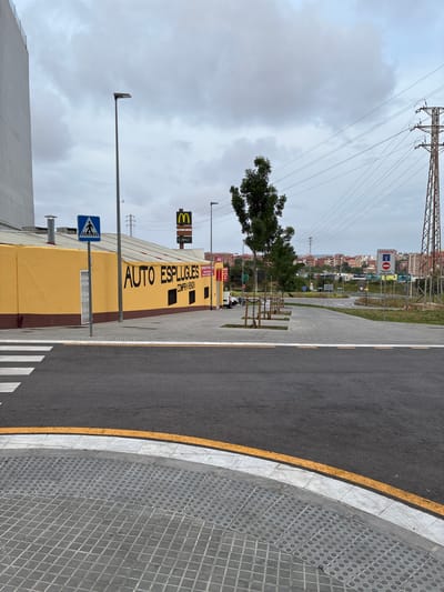 The image captures an outdoor urban street scene in Esplugues de Llobregat, Spain, under an overcast sky during daytime. The perspective is tilted, showing a road, sidewalk, and various structures.
In the immediate foreground, a dark asphalt road with a prominent yellow line and a white line defining its edge curves gently to the right. Adjacent to the road is a grey tiled sidewalk, featuring both square and circular patterned paving stones. A white zebra-striped pedestrian crossing is partially visible in the lower left.
The mid-ground is dominated by a long, low yellow building with black text painted on its side, reading "AUTO ESPLUGUES" and "COMPRA VENTA" (Buy Sell). A blue square sign with a white pedestrian crossing symbol is mounted on a pole directly in front of this building. Above the building, a yellow McDonald's 'M' logo is visible on a tall signpost. Further down the street, a red and white traffic sign displays "ATENCIO" (Attention), a "No Entry" symbol ("SENTIT PROHIBIT"), and "EXCEPTE ACCES MAU" (Except Maintenance Access). A white van is parked near the building's entrance, which has a partially visible red sign marked "ENTRADA" (Entrance).
To the right of the yellow building, a paved pathway lines a grassy strip with small, sparsely leafed trees. In the background, numerous residential buildings with varied colors can be seen on a distant hillside. Overhead, a complex network of power lines stretches across the cloudy grey sky, supported by a large metal electricity pylon on the left. The overall impression is one of a functional commercial/industrial area bordering a residential zone.