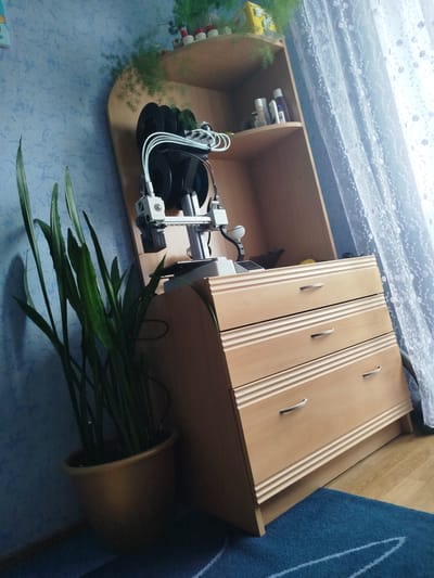 In an indoor setting, possibly a bedroom or office, a wooden chest of drawers with three drawers is positioned next to a wall with blue textured wallpaper. On top of the chest of drawers, a 3D printer is visible, with spools of black filament loaded onto it. To the left of the chest of drawers, a tall green plant in a gold pot stands on the floor. Above the 3D printer, a wooden shelf unit holds a few items, including bottles and greenery. A light-colored sheer curtain with a floral pattern is to the right of the scene, and a blue rug is on the floor in front of the chest of drawers. The image appears to be taken in Vynnyky, Ukraine, although this cannot be confirmed from the image alone.