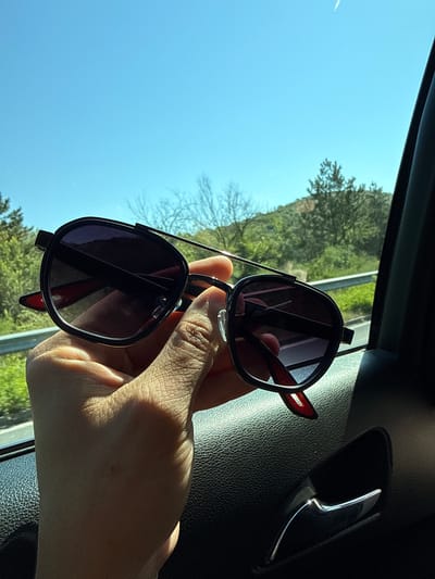 A hand holds a pair of black aviator sunglasses with red accents in front of a blurred outdoor scene of green hills and trees under a clear blue sky. The sunglasses have dark lenses and a double bridge. The hand and sunglasses are in focus, while the background is out of focus, suggesting the photo was taken from inside a vehicle. The interior of the vehicle, with a black textured surface and a chrome door handle, is visible in the foreground and to the right. The image does not provide any information about the city of Bliznets, Bulgaria.