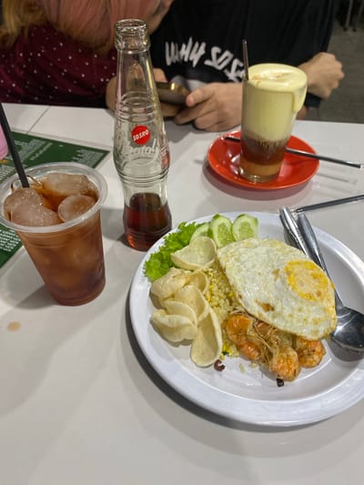 On a white table, a meal is served. A plate of fried rice, topped with a fried egg and three shrimp, sits next to a glass of iced tea and a bottle of Sosro tea. Another glass, containing a layered beverage with a frothy top, rests on a red saucer. In the background, two people are visible, one wearing a maroon shirt with white polka dots and the other in a black t-shirt. The scene appears to be in a dining establishment, possibly a restaurant in Mekar Bakti, Indonesia, given the presence of local brands like Sosro.