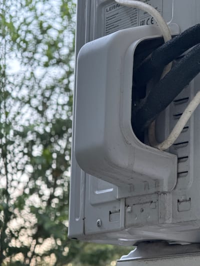 The image shows a close-up of the exterior unit of an air conditioning system, likely installed on the side of a building. The unit is white and shows signs of weathering and dirt. Two insulated pipes, one black and one white, exit the unit through a cutout in the casing. A label with text and the "CE" mark is visible on the unit. The background consists of blurred green foliage, suggesting an outdoor setting possibly in a residential or semi-urban area. There are no people or animals visible in the image. The focus is on the details of the air conditioning unit and its connections.