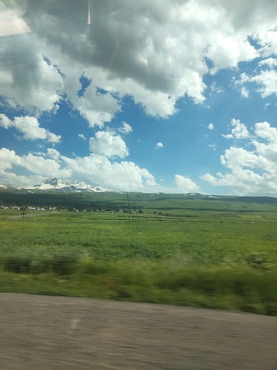 The image depicts a scenic landscape under a partly cloudy sky. The foreground is dominated by a blur of green fields, suggesting motion, possibly from a vehicle. This blur transitions into a more defined middle ground featuring more green fields, dotted with what appear to be small buildings and trees, forming a village or rural settlement. In the background, a range of mountains stands tall, their peaks capped with snow, indicating a cooler climate or high altitude. The sky is a vibrant blue, with large, fluffy white and grey clouds scattered across it, suggesting fair weather with some cloud cover, possibly late morning or early afternoon due to the brightness. The overall impression is one of natural beauty and tranquility, with the snow-capped mountains providing a dramatic backdrop to the verdant plains and the distant settlement. No people or specific objects are clearly discernible in the image, other than the general landscape features. There is no visible text in the image.