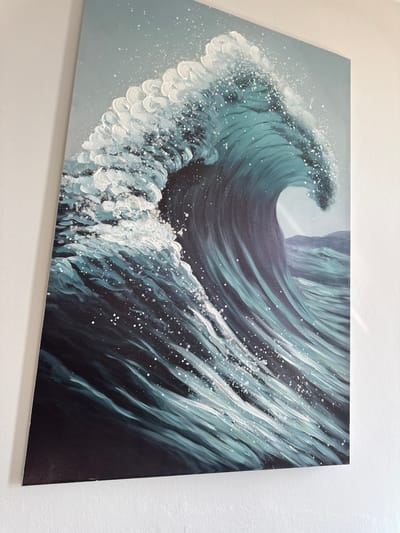 The image shows a close-up of a painting of a wave. The wave is depicted in shades of deep blue, teal, and white, with white spray and bubbles scattered across its surface. The painting is hanging on a white wall. There is no indication of a specific location in Bashkia Durrës, Albania.