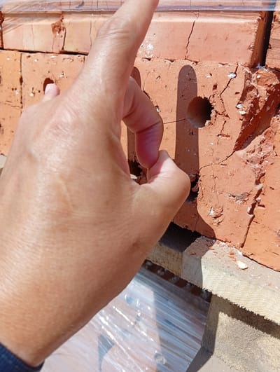 A close-up shot captures a person's hand touching a cracked red brick. The hand, with its knuckles prominent, is positioned in the foreground, obscuring a portion of the brick. The brick itself shows visible fractures and a circular hole, a common feature in some types of bricks used for construction. The background reveals more stacked bricks, suggesting a construction or storage area. The overall scene is outdoors, with sunlight casting shadows on the bricks. The location is identified as Қызылжар ауылдық округі, Kazakhstan.