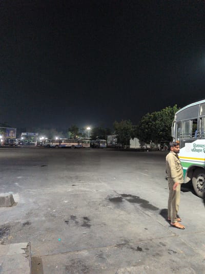 The image depicts a dimly lit outdoor scene at what appears to be a bus depot in Kajheri, India. The sky is dark, indicating nighttime or very early morning. In the foreground, a man dressed in a light brown uniform and sandals stands to the right of the frame, looking towards the left. He is positioned next to a white and green bus with text in Hindi on its side, partially visible. The bus is large and appears to be a public transport vehicle.

In the background, several other buses are parked in rows. Some of these buses are also white and blue. A structure resembling a bus shelter or canopy with lighting is visible behind the parked buses. Further in the distance, there are trees and some buildings. The ground is paved with asphalt and shows some wet patches and debris. A large poster with an image of a person in traditional attire is also visible on a billboard to the left, near some shops. The overall impression is of a quiet bus station during off-peak hours.