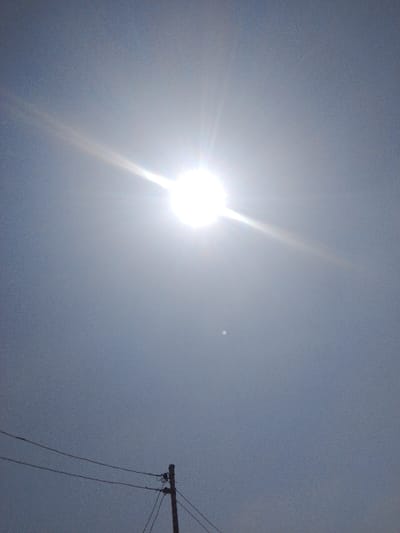 The scene depicts a bright, clear daytime sky in Okinni, Nigeria. A brilliant white sun, emitting strong diagonal lens flares, is positioned in the upper-left quadrant of the light blue to greyish-blue sky. A faint, small, white circular object, consistent with the moon, is visible below and to the right of the sun. In the lower portion of the frame, a segment of a dark brown utility pole is present, from which multiple black electrical or communication wires extend horizontally across the lower third of the image. No individuals, specific actions, or interactions are observable.