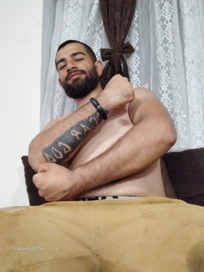 A single adult male is depicted from the waist up, seated indoors in what is identified as Oreshene, Bulgaria. The subject, appearing to be in his 20s or 30s, has dark hair, a beard, and a mustache, and is looking directly at the camera with a subtle expression. He is shirtless, revealing his chest and arms. A prominent dark-inked tattoo is visible on his left forearm, consisting of block letters spelling "GOD" or similar, reversed relative to the viewer. A dark, possibly braided, bracelet is worn on his left wrist. His left arm is bent at the elbow, with the forearm raised and the fist clenched near his chest. His right arm is also bent, resting against his torso. He wears light-colored underwear with a visible pattern along the waistband, above tan or light brown trousers that occupy the lower portion of the frame. The background features a plain light-colored wall to the left and layered curtains to the right. The inner curtain is white, sheer, and patterned, while an outer dark brown curtain is gathered and tied with a bow-like knot in the center. A dark brown cushion or part of a couch is visible behind the subject. The image displays a "Galaxy A05s" watermark in the lower-left corner.