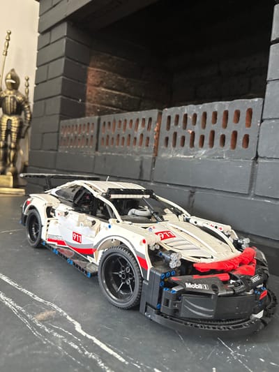 In Хохряки, Russia, a LEGO Technic model of a white Porsche 911 race car with red and black accents is displayed on a dark surface, possibly a table. The car features a prominent rear spoiler, detailed wheels, and racing decals including "911" and "Mobil 1". To the left of the car, a vintage-style metal figurine of a knight in armor holding a spear stands against a dark brick fireplace. The fireplace itself is constructed from dark grey bricks with an opening at the top and a grate-like structure in front of it, revealing reddish-brown bricks behind. The lighting in the scene is somewhat dim, casting shadows and highlighting the details of the car and the knight.