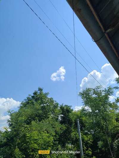 This is a daytime, eye-level, outdoor photograph taken in Moldoveanca, Moldova. The scene is dominated by a bright blue sky with scattered white clouds and a portion of a roof structure in the upper right corner. Several dark power lines traverse the sky. Below the sky, lush green trees fill the lower half of the frame, with a concrete utility pole visible on the right side of the image. The bottom of the image contains a watermark indicating "realme Shot on GT Master." No people or specific actions are depicted.