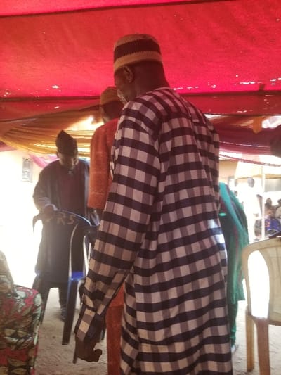 This image depicts an outdoor gathering in Ochadam, Nigeria, under a temporary shelter. A prominent red fabric canopy covers the overhead space, with a lighter, possibly yellow, fabric draped decoratively beneath it. In the immediate foreground, an adult male stands with his back to the camera, wearing a long-sleeved, loose-fitting garment with a black and white checkerboard pattern and a striped knit cap. Directly behind him, another adult male, also seen from the back, wears a bright orange traditional garment and a cap. Further to the left in the mid-ground, a third adult male, dressed in a dark blue or black garment and a dark cap, is observed interacting with a black plastic chair that bears white "RS" lettering on its backrest. A light-colored plastic chair is partially visible on the far right, and a section of a patterned armchair with red, black, and white designs is present in the lower-left corner. The ground appears to be unpaved, consistent with dirt or packed earth. Blurred figures populate the background, indicating the presence of additional attendees. The scene is illuminated by bright natural light.