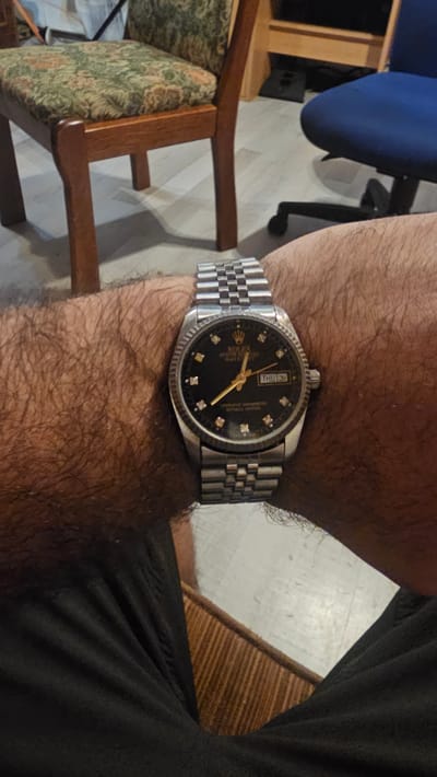 In Oelsberg, Germany, an individual wearing a black t-shirt and a Rolex Datejust watch with a black dial and diamond hour markers is seated. The watch displays the time as 4:23 and the date as Thursday, the 31st. The background shows a wooden chair with a floral pattern fabric seat, a blue office chair, and a tiled floor.