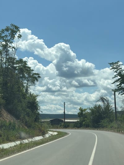 This is a vertical, eye-level, medium shot of a curved asphalt road with a white lane divider, heading into the distance. The road is flanked by lush green vegetation on both sides. In the foreground, a concrete ditch runs along the left side of the road. In the background, there are distant fields and a building with a dark roof. The sky above is a vibrant blue, filled with large, fluffy white cumulus clouds. Two wooden utility poles with wires are visible in the middle ground. The overall scene depicts a rural roadside in Garchinovo, Bulgaria, on a bright, cloudy day. There are no people or animals visible in the image.