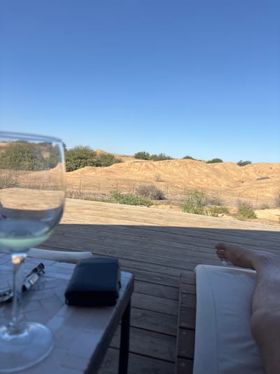The image depicts an outdoor leisure scene in an arid environment, consistent with the Merhavim Regional Council area in Israel. In the foreground, a person's bare right leg and foot are visible, resting on a white cushion of a lounge chair positioned on a wooden deck. Adjacent to the person, on a small table with a light-colored tiled surface, a clear wine glass partially filled with liquid stands next to a dark, rectangular wallet and a metallic object. A prominent shadow line runs diagonally across the wooden deck in the midground. The background features undulating, light brown, sandy hills and dry terrain, sparsely covered with green and dry scrub vegetation and small trees. A wire fence is faintly discernible at the base of the hills. The sky above is clear and blue.