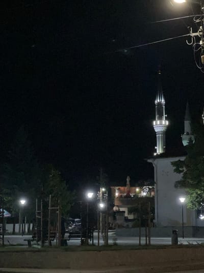 The image displays a nighttime scene in Kornitsa, Bulgaria, featuring an illuminated mosque and a public park area. On the right, a prominently lit minaret of a mosque stands tall, displaying white and possibly bluish-white illumination on its balconies and spire. A second, darker minaret is partially visible behind it. The mosque's main structure, light-colored, is partially visible below the illuminated minaret. To the left of the mosque, a multi-story building is illuminated, revealing windows and a rooftop terrace with at least two open umbrellas and additional lighting. The foreground and left side of the image show a paved park or plaza with several modern streetlights emitting white light, illuminating young trees supported by wooden stakes, and at least two park benches. A dark trash receptacle is visible on the right edge of the park area. In the mid-ground, a dark vehicle, possibly an SUV, is parked or moving on a street behind the park. Two indistinct human figures are vaguely discernible: one near the young trees on the left and another near the parked vehicle. Overhead power lines and utility poles with attached wires are visible against the dark night sky in the upper right quadrant. The scene is artificially lit, creating areas of bright illumination contrasted with deep shadows, and exhibits digital noise characteristic of low-light photography.