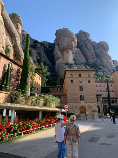 The image shows the Montserrat Monastery, located in Monistrol de Montserrat, Spain, on a clear day. In the foreground, two women are standing and looking away from the camera. One woman is wearing a white hat and a white top with blue pants, and the other is wearing a patterned shirt and striped pants. Behind them, a wall of red roses is visible, and beyond that, a building with an orange facade and a tall, slender evergreen tree. Further in the background, a large, multi-story brick building with numerous windows stands against a backdrop of imposing, rugged rock formations. Several other people are visible in the distance, including one taking a photograph. A bicycle traffic sign is also present. The sun is casting shadows, indicating it is likely morning or afternoon.