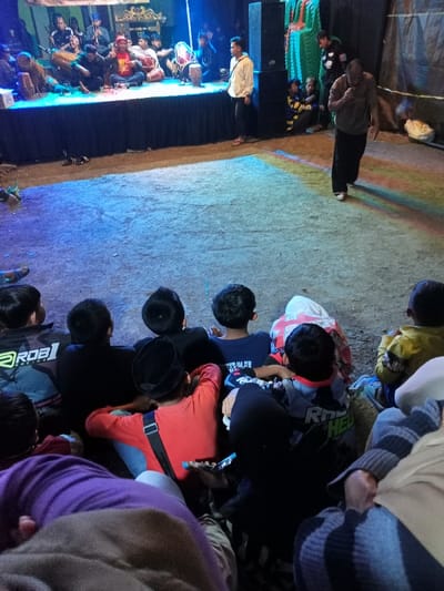 This nighttime image captures a traditional music performance in Pangalengan, Indonesia. A group of musicians is seated on a raised stage draped with black fabric in the background, actively playing various instruments, most notably several kendangs (traditional drums). One musician in a red patterned top is prominently visible. Large speakers and stage lights emitting vibrant blue and green hues illuminate the performance area, casting colorful patches onto the dirt ground below. A man in a white shirt stands near a speaker on the stage.
In the midground, an adult male dressed in a dark shirt and dark pants walks across the illuminated dirt field, away from the stage. The foreground is filled with an audience of mostly children and some adults, seen from behind, sitting on the ground and intently watching the performance. Several children wear hats (kufi-style), and one child's black and white shirt has "ROB" visible on the back. Another's shirt shows "RCH HEA" partially. The setting appears to be an informal outdoor venue, suggesting a local community event or cultural gathering. The atmosphere is one of engagement and observation, with the audience focused on the stage.