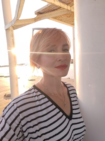 A medium shot photo shows a blonde woman with auburn hair, wearing a black and white striped V-neck top and a necklace. She is standing outdoors near a white pillar and a metal structure, with the sun in the background, casting lens flare. The woman is looking toward the camera with red lipstick. The scene appears to be a pier or boardwalk.