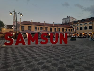 The image depicts the "Samsun" sign in a public square in Samsun, Turkey, during twilight. The sign, made of large red letters, is positioned in the foreground on a patterned paved surface. Behind the sign, a streetlamp with two lit bulbs is visible, along with banners displaying fashion models. Further back, a row of buildings with orange-tiled roofs and arched windows lines the square. Some buildings have awnings extended over storefronts, and several people are seated at outdoor tables. A modern multi-story building stands in the background to the left, and a minaret is also visible. The sky is overcast, and the overall lighting suggests the transition from day to evening. Several individuals are walking in the square, some appearing to be patrons of the establishments.