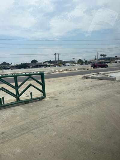 The image, taken from a slightly elevated perspective, is rotated 90 degrees counter-clockwise, showing a roadside view under a partly cloudy sky during daylight hours.
In the foreground, on the right side, there is a rough, concrete or asphalt surface, possibly a sidewalk or shoulder, exhibiting tire marks and scuffing. A prominent green metal barrier or fence structure with angular patterns occupies the lower right section of the foreground. Reflections of what appear to be interior vehicle components or structures are visible in the upper left corner, suggesting the photo was taken from inside a vehicle.
Moving towards the midground, a paved road or street is visible, with concrete curbstones or barriers separating it from the foreground area. Several vehicles are parked or moving along the road. A dark red SUV is distinctly visible, and other lighter-colored cars can be seen further down the street. Utility poles with multiple power lines or cables stretch across the frame, running parallel to the road against the sky.
In the background, residential or commercial buildings with dark roofs are partially obscured by trees and other vegetation. Some palm trees are identifiable among the greenery. The sky above is a light blue with scattered white clouds. The overall scene depicts a typical street environment in what appears to be an urban or suburban setting. No specific activities or events are actively taking place, and no visible text can be discerned in the image.