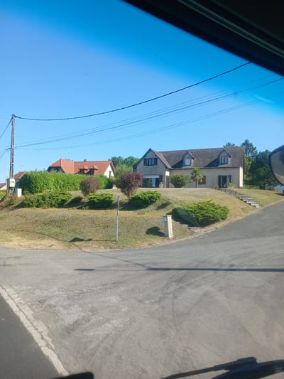 This image captures a suburban scene in Cléry-sur-Somme, France, on a clear day. The foreground shows a paved road with white markings. Beyond the road, a gently sloping, grassy embankment is dotted with green bushes and shrubs. Several houses are visible on the embankment. One house on the left has a red tiled roof and is partially obscured by dense green hedges. To its right, a larger house with a grey tiled roof and beige walls stands on a raised lawn, accessible by a set of concrete stairs. This house features dormer windows on its roof and several windows on its facade, with brown shutters on some. In the background, a line of trees forms a natural boundary. A utility pole with wires is visible on the left side of the image, and a small mailbox-like structure sits on the embankment near the road. The sky is a vibrant blue.