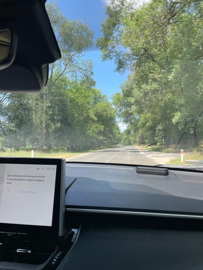 The image shows a view from inside a car driving on a paved road lined with trees. The road appears to be in a rural area, possibly in Coșernița, Moldova, given the context. The sky is blue with some clouds, and the sun is shining. The car has a digital display in the foreground showing text in Russian which translates to "To save all personal settings in this car, create a profile below." Below this text, there is a button labeled "Create profile". The car's interior, including the dashboard and part of the windshield, is visible. There are no people visible in the image. The scene depicts a typical daytime car journey through a tree-lined road.