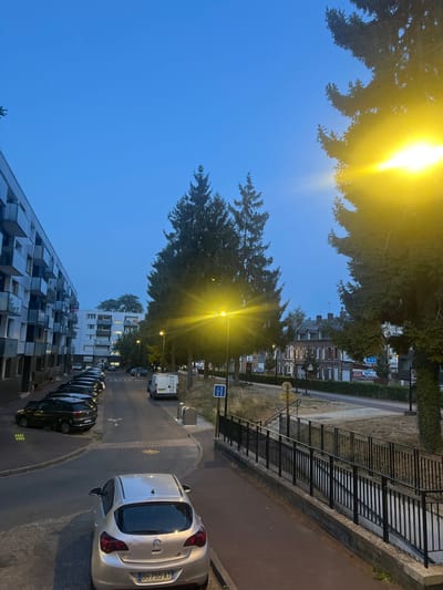 This is an outdoor, eye-level, vertical, full shot of a street scene in Maromme, France, captured during twilight. The sky is a clear, deep blue, with a bright yellow street lamp illuminating the right side of the frame, its light streaking through the branches of a tall pine tree. On the left, a modern, multi-story apartment building with balconies lines the street. A row of parked cars is visible along the curb, predominantly dark-colored sedans and hatchbacks. A silver Opel Astra hatchback is parked in the foreground, facing away from the camera, its license plate readable as "BH-755-AT". A white van is parked further down the street, near a blue and white pedestrian crossing sign. Across the street, a row of brick buildings with pitched roofs can be seen. A black metal railing lines a raised walkway or path running parallel to the street, with a set of stairs descending from it. The ground is paved with asphalt and cobblestones. The overall scene suggests a quiet residential neighborhood at dusk.