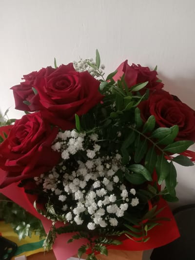 The image shows a bouquet of red roses and baby's breath flowers, wrapped in red paper. The bouquet is set against a white wall and on a wooden surface. There are also some green leaves and a black object, possibly a phone, visible at the bottom left of the image, along with a colorful fabric that resembles a flag. The scene appears to be indoors, and given the presence of indoor objects and the white wall, it is likely a domestic or indoor setting. The specific location within the city of Kielce, Poland, cannot be determined from the visual information provided.
